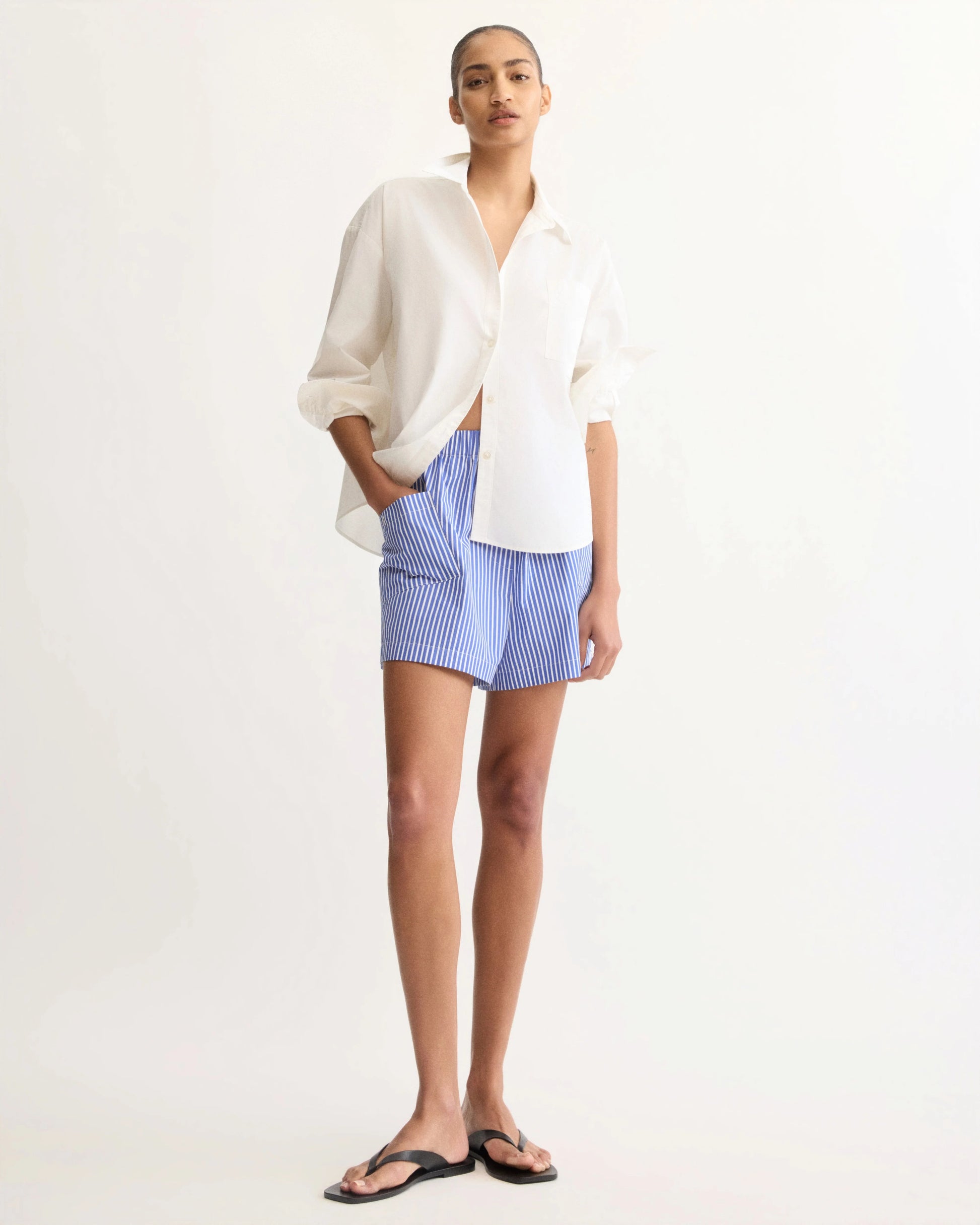 Model wearing white oversized button-up shirt with blue striped shorts and black sandals