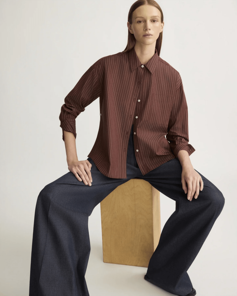 Model wearing oxblood striped button shirt with wide-leg navy trousers seated on wood block